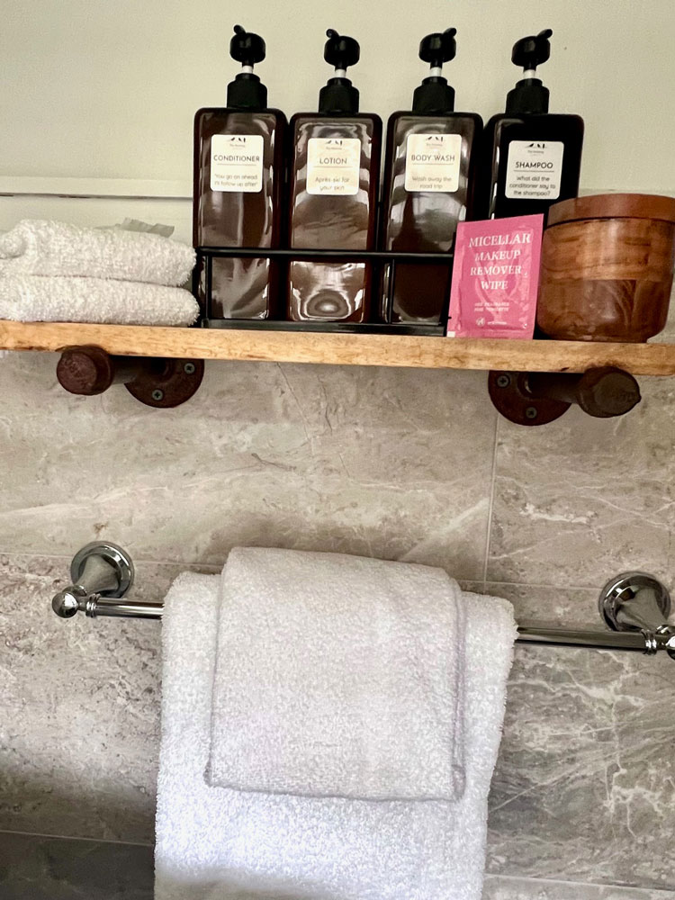 A bathroom shelf in this Accessible Lodging Wilmington VT holds bottles of conditioner, lotion, body wash, and shampoo, along with folded white towels, a pink packet of wipes, and a wooden container. More white towels hang on a rack below.