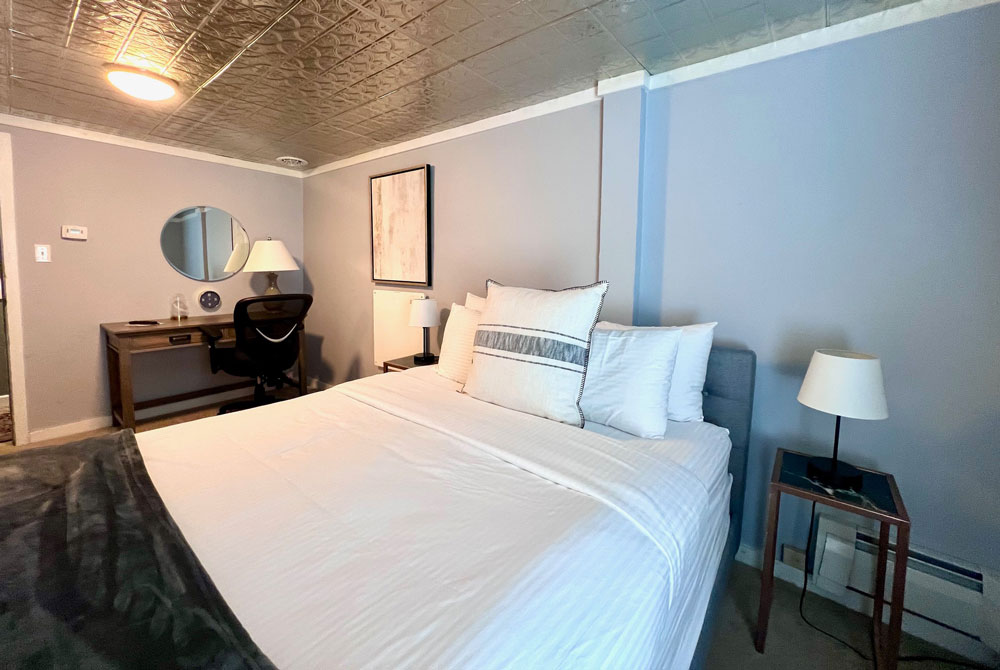 A cozy bedroom in accessible lodging, Wilmington VT, featuring a large neatly made bed with white bedding, blue-gray walls, a side table with lamp, and a desk with chair and round mirror. The ceiling has a unique metallic patterned finish.