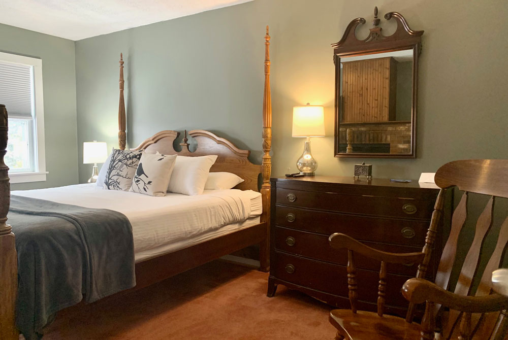 A cozy bedroom reminiscent of boutique inn rooms in Vermont, featuring a wooden four-poster bed, white bedding, decorative pillows, a gray throw, dark dresser with mirror, table lamp, rocking chair on brown carpet, and natural light from a window.